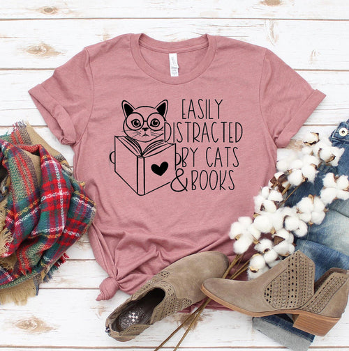 Pink t-shirt with cat and book design, surrounded by boots, plaid fabric, and cotton on a wooden surface.