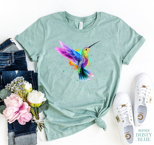 Mint green t-shirt with colorful hummingbird design, paired with jeans, sneakers, and flowers on a white background.