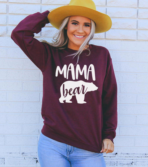 Woman wearing a maroon 'Mama bear' sweatshirt against a light brick wall.
